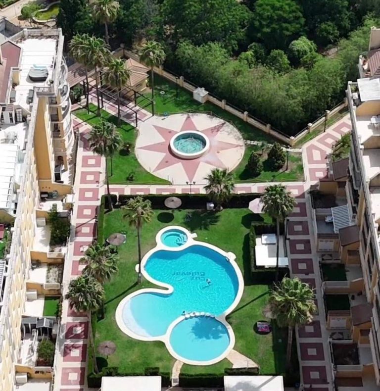 Piscina comunitaria en la urbanización del apartamento, rodeada de jardines y césped, ideal para disfrutar del sol en Denia